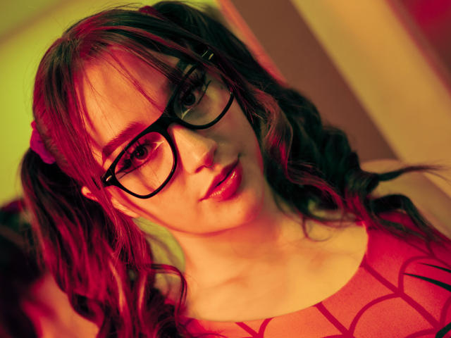 CristySanz with red-tinted hair in pigtails, wearing black glasses and a red top with geometric patterns, has a cute face and slightly parted lips, looking directly at camera.