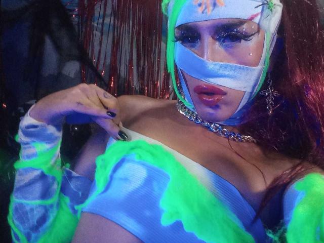 Crystalinn with neon green and blue glow-up, wearing a mask and chain, showing her tits in a glow-in-the-dark top, black nail polish, and a cross earring.