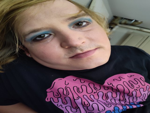 Ava Brayns met blauwe oogmake-up, blond haar en een roze T-shirt met een hartprint, kijkt omhoog met een lichte grijns en laat een kleine neusring zien..