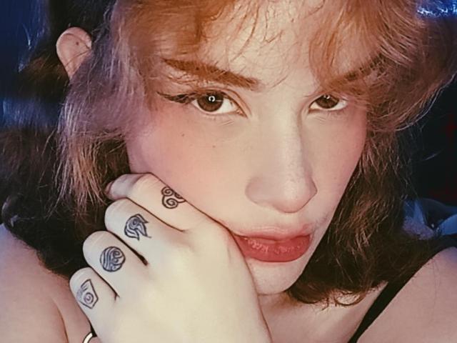 DanaStone has red-brown hair, brown eyes, and tattoos on her fingers with symbols like eyes and hearts. She's biting her finger with pink lips, looking at the camera.