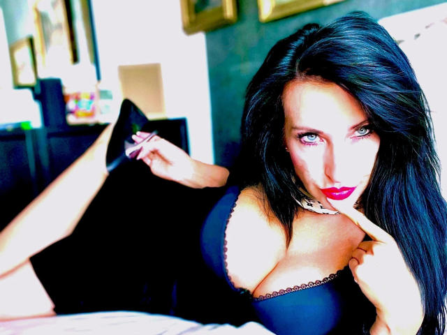 Lady4youLive-hot with dark hair, red lipstick, and black lingerie, lying on bed, touching her lips, showing large tits, black choker, and black high heels, looking seductively at camera.