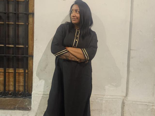 Nahirr stands with arms crossed, wearing a black dress with gold stripes, looking to the side with a slight smile, tattoo on forearm, black hair, white wall behind.
