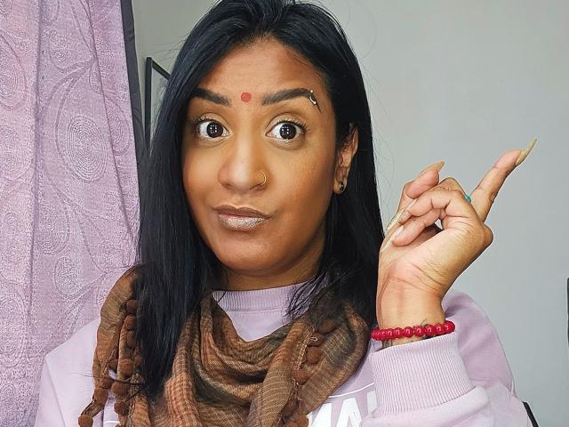 Nahirr has brown skin, black hair, and a red bindi on her forehead. She's wearing a pink shirt and brown scarf, with a red bead bracelet. She's making a rock gesture.