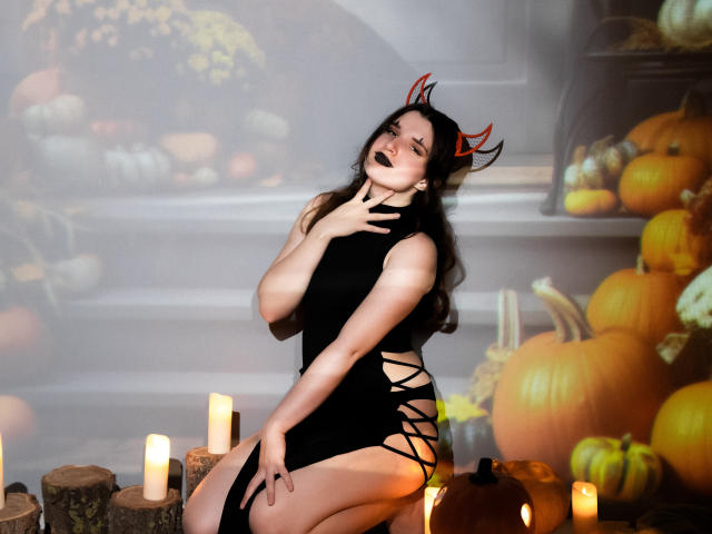 BonnieFlower con maquillaje oscuro y cuernos rojos de diablo, vestida con un vestido negro con cordones, arrodillada entre velas y calabazas, tocándose el cuello, mostrando su trasero y con lápiz labial negro..