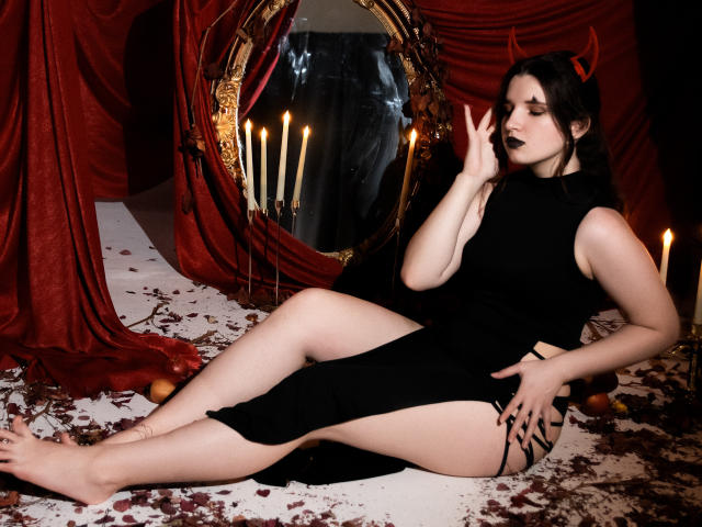 BonnieFlower with pale skin, black dress, red devil horns, dark lipstick, sitting on bloody white floor, black skirt lifted, hand touching face, surrounded by candles and red drapes.