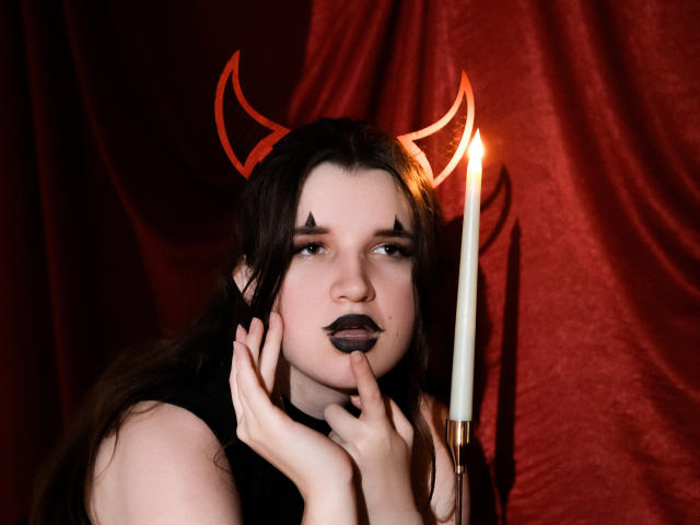 BonnieFlower with dark makeup, devil horns, and black lipstick, hands on face, holding a candle, red velvet background, pale skin, long brown hair, serious expression, soft lighting.