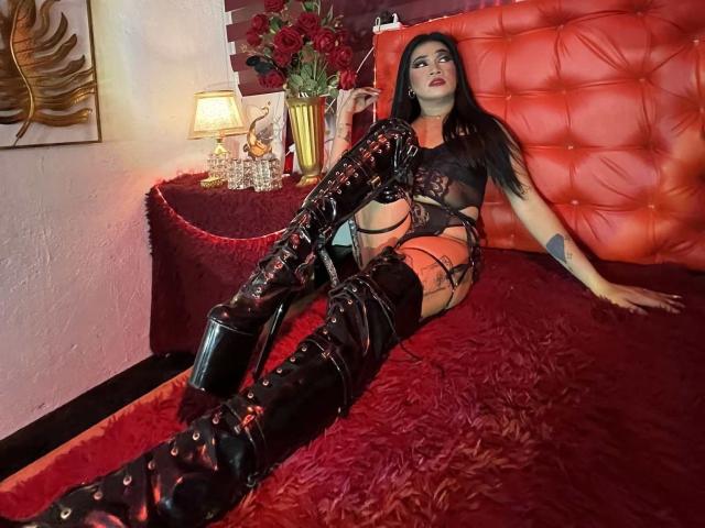 Séduction asiatique avec lingerie noire en dentelle, bottes noires brillantes montantes, et de longs cheveux noirs, allongée sur un lit rouge avec une tête de lit rouge capitonnée, regardant de manière séduisante vers le haut..