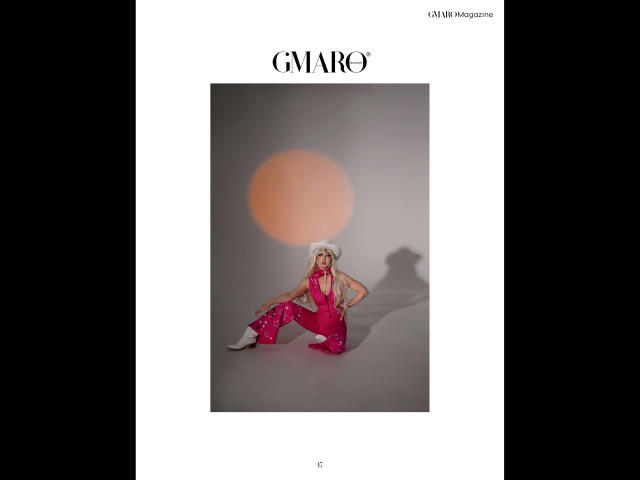 Shezel, con un sombrero blanco, pantalones rosa brillantes y una blusa roja, sentada con las manos en las rodillas, con la sombra de un círculo similar al sol en una pared gris, y zapatillas blancas..