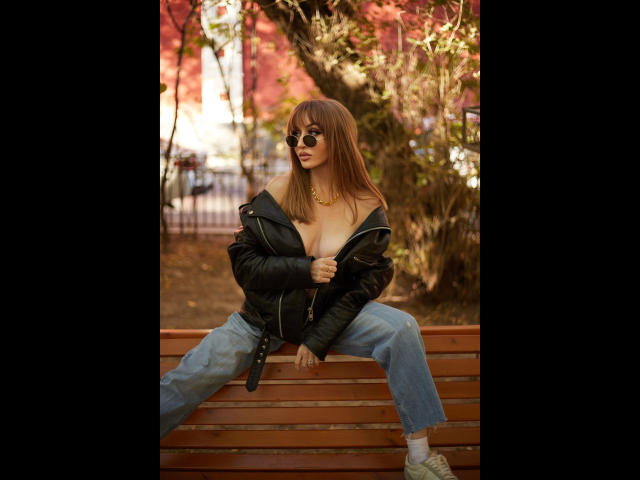 Shezel, avec ses cheveux bruns, portant des lunettes de soleil, une veste noire et un jean bleu, assise sur un banc en bois, montrant ses seins, avec un collier en or et des baskets blanches..