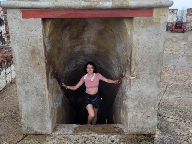 GabrielaMoore standing in a dirty, stone, tunnel-like structure with her hands on the walls, wearing a pink crop top and black shorts, smiling with her legs apart.