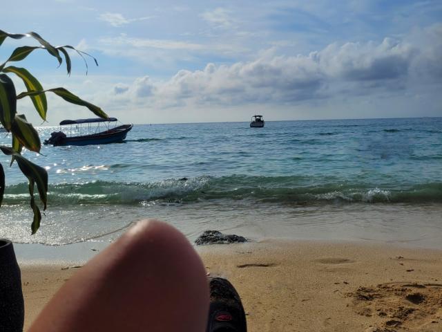 GabrielaMoore's leg in the foreground, blue-green ocean, small boat on left, bigger boat in center, leafy branch on left, sandy beach, cloudy sky, calm water.