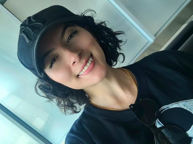 GabrielaMoore with curly black hair, wearing a black baseball cap and black hoodie, smiling with white teeth, brown eyes, gold chain, and sunglasses hanging from her hoodie.