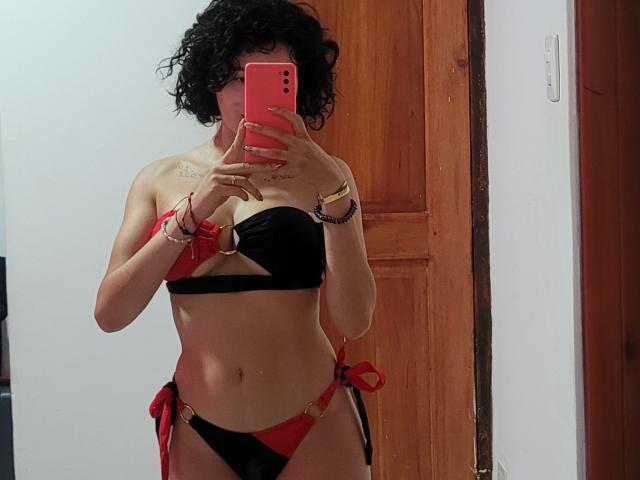 GabrielaMoore with curly black hair, wearing a red and black bikini, holding a pink phone, showing her slim, tan body, small tits, and a tattoo above her chest.