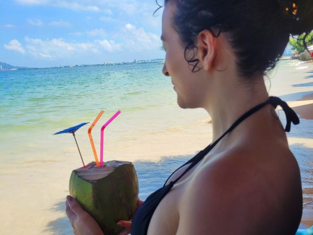 GabrielaMoore with wet black hair, holding a green coconut with pink and orange straws and a small blue umbrella, standing on a sandy beach, wearing a black bikini top.