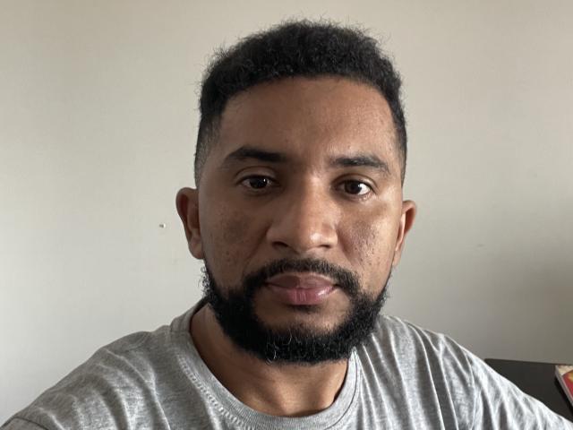 SilverHotMan has a short, curly black beard, brown eyes, and a light brown skin tone, wearing a gray shirt, looking serious with slightly parted lips.