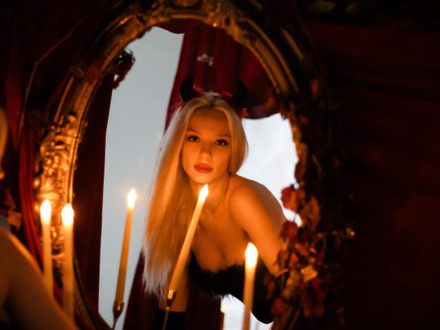 LexyGlam with long blonde hair, black devil horns, and black lingerie, leaning into an ornate mirror, holding a lit candle, her tits visible, surrounded by more candles.