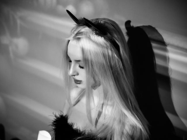 Black-and-white photo of LexyGlam with long blonde hair, wearing a feathery headband and fur collar, eyes closed, shadow on the right side of her face.