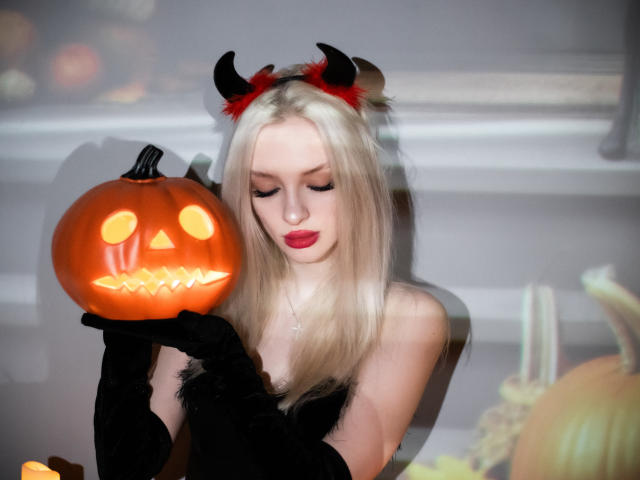 LexyGlam with blonde hair, red devil horns, black gloves, holding a carved pumpkin with a spooky face, pale skin, red lipstick, and a black strapless top.