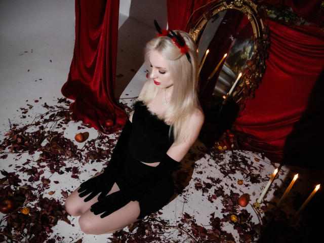 LexyGlam mit heller Haut und blonden Haaren, trägt schwarze Teufelshörner, ein Kleid und Handschuhe, kniet auf einem weißen Boden mit roten Blättern, schwarzen und roten Kerzen und einem verzierten Spiegel..