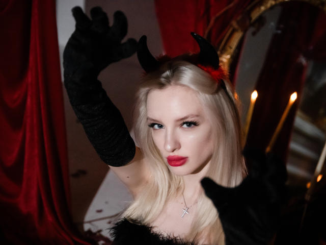 LexyGlam con i capelli biondi, corna da diavolo nere, rossetto rosso, guanti neri e una collana con una croce, che alza le braccia e guarda seducentemente la fotocamera. Il seno è visibile sotto un top nero senza spalline..