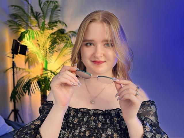 BonnieBell with blonde hair, blue eyes, and a slight smile, holding glasses in front of her chest, wearing a black floral off-shoulder top, and a silver necklace.