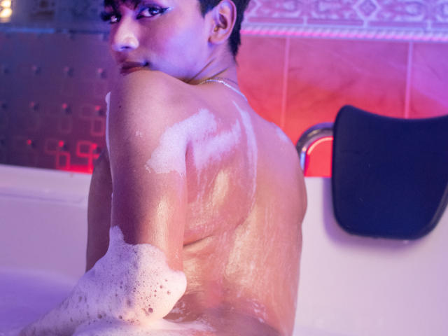 Anddyys with soapy tits, back to the camera, in a bathtub. Purple and pink lights reflect on their wet skin. Black bath pillow on the right. Short, dark hair.