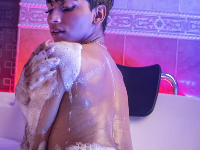 Anddyys is in a bathtub, covered in soap suds, with a black chair in the background. They have short brown hair, a gold necklace, and a sexy, focused expression.