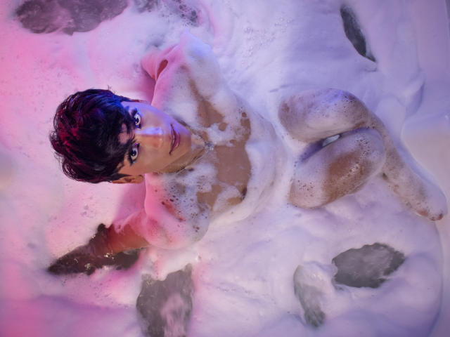 Anddyys is lying in a snow bath, surrounded by foam, with pink and purple lighting. Anddyys has short black hair, is nude, with visible breasts and a cute face.