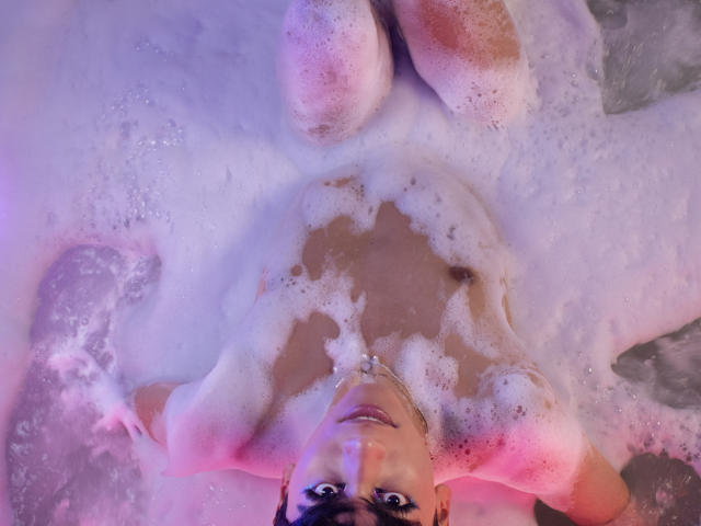 Anddyys is in a bubble bath, her tits and ass partially covered in white foam, with pink and purple lighting. She's lying on her back, looking up.