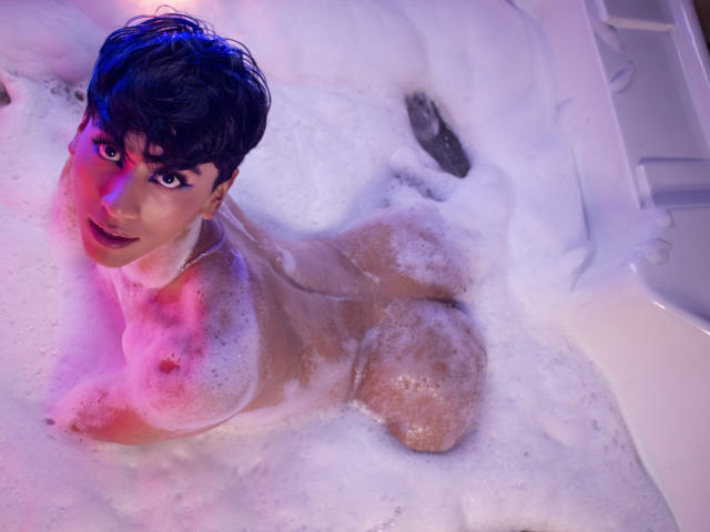 Anddyys with short black hair, nude, in a bubbly white bathtub, lying on their side, showing their tits and ass, with purple and pink lighting on their face and chest.