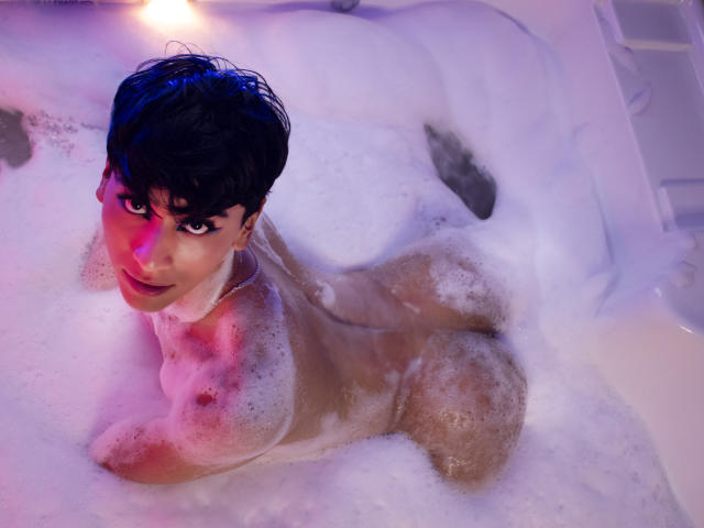 Anddyys with short black hair, nude, in a bubbly white bath, lying on her side, showing her ass and tits, with a purple and pink light on her face.