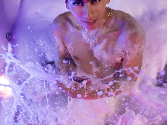 Anddyys in a purple-lit bathtub, naked with soap suds covering his chest and arms, splashing water with a focused, slightly sweaty expression, wearing a gold necklace.