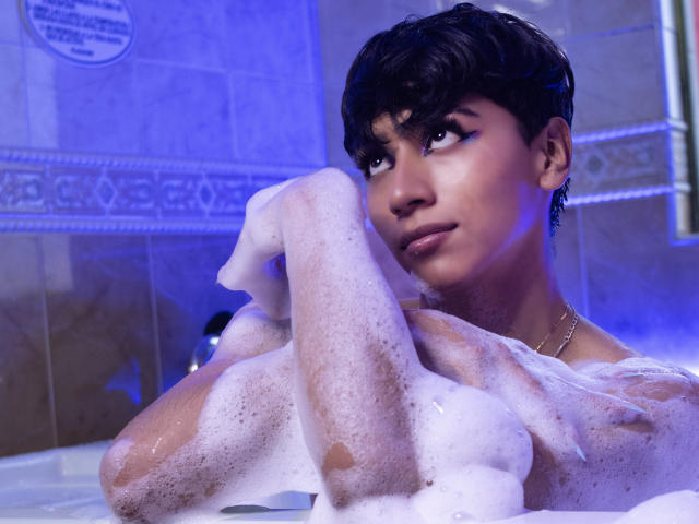 Anddyys with short black hair, soap suds on arms and chest, looking up, blue light, dark eyes, subtle smile, earrings, necklace, and soft skin. Bath scene.