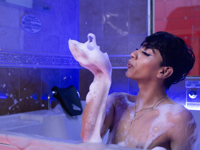 Anddyys with short black hair, soap suds on arm and chest, lifting arm to face, blue bathroom light, wet skin, necklace, looking up, slightly open mouth.