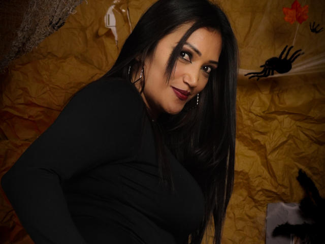DayanaMarians69 met lang zwart haar, gekleed in een zwarte top met lange mouwen, kijkt over haar schouder, glimlachend, rode lippenstift, oorringen, bruine ogen, liggend op gele, gekreukelde stof..