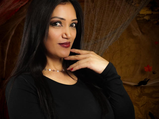 DayanaMarians69 with long black hair, dark red lipstick, and a black long-sleeve top, hand near face, wearing a pearl necklace, looking at camera with a slight smile.