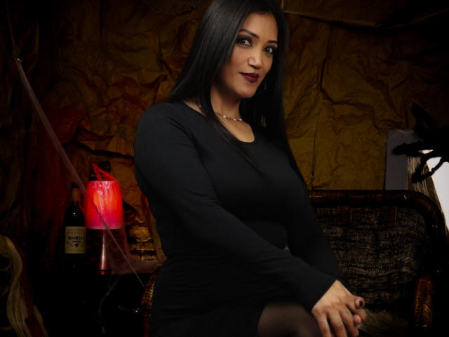 DayanaMarians69 with long black hair, wearing a tight black dress, sitting, showing her ass, a necklace, and a confident smile, with a red lamp and bottle in the background.