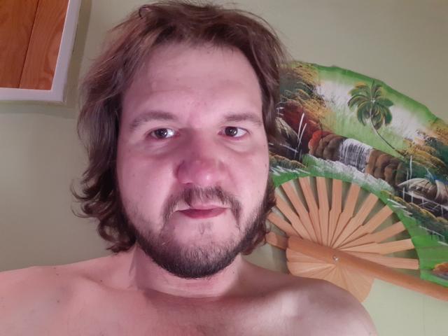 NectarineFad has a chubby face, brown wavy hair, and a beard. He's shirtless with a neutral expression and a small nose. His tits are not visible.