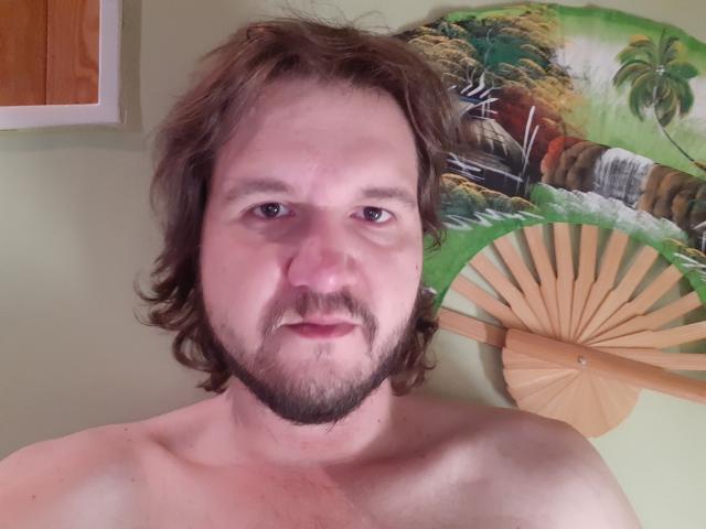 NectarineFad has a chubby face with brown wavy hair, a beard, and a mustache, shirtless with a pale skin tone, standing in front of a wooden fan.