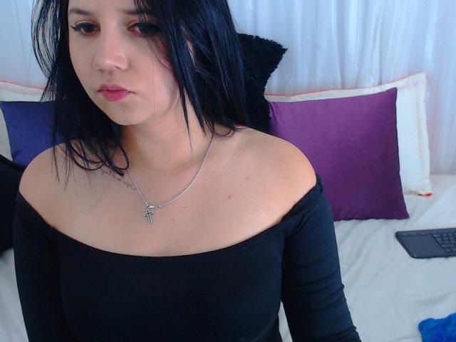 Kkatalina met zwart haar, bleke huid, gekleed in een zwarte top met open schouder, een zilveren ketting met een kruis, en subtiele make-up, staat voor een bed met paarse kussens..