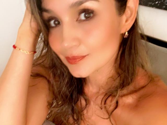 SofiaVerlek's face, brown wavy hair, subtle makeup, red lipstick, small gold earrings, red bracelet, slight smile, looking at camera, right hand touching head, tan skin, attractive.