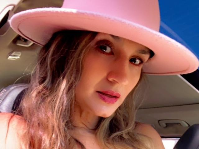 SofiaVerlek with pink hat, brown wavy hair, and pink lips, sitting in a car, looking at camera, wearing no top, showing bare shoulders, with blue sky outside.