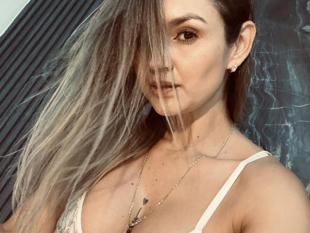 SofiaVerlek has long blonde hair covering one eye, wearing a white spaghetti strap top, showing small heart tattoo on chest, and a necklace with a small pendant.