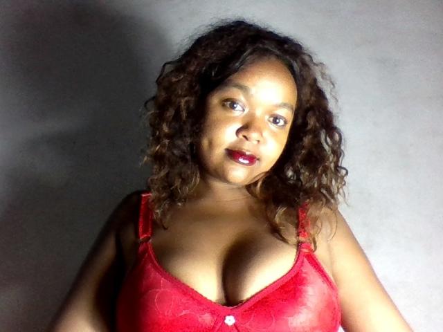 LoreineHot with curly brown hair, red lipstick, and red lace bra, showing her big tits against a gray wall, looking slightly up with a soft smile.