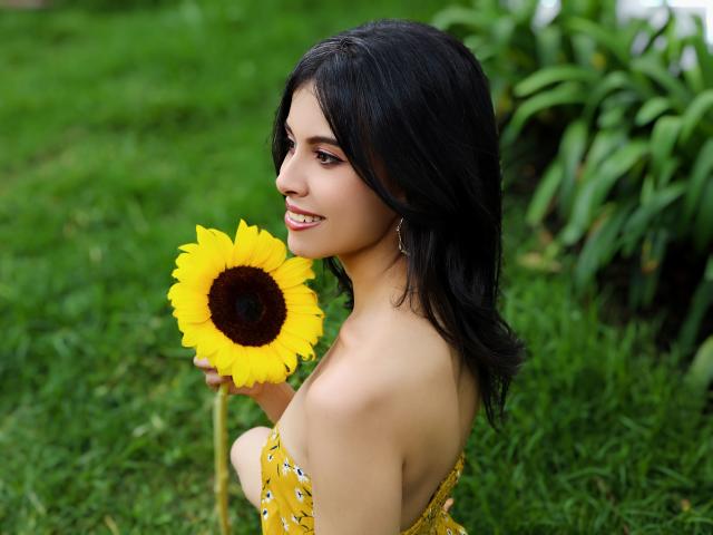 TaylaCooperk, avec ses cheveux foncés, tenant un grand tournesol jaune, souriante, portant une robe jaune à fleurs, montrant son dos et son épaule nue, debout sur de l'herbe verte..