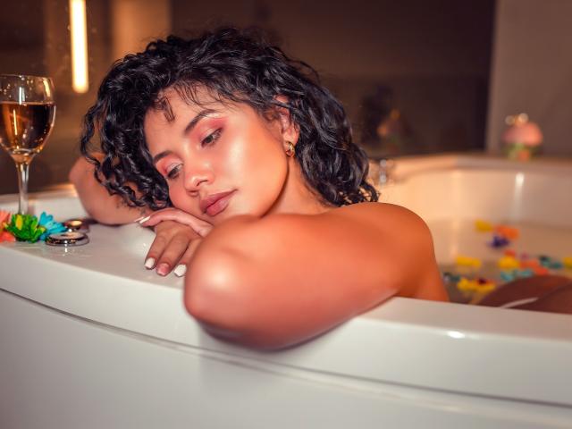 CanelaLebrad with curly black hair, pink eyeshadow, and gold earrings, leans on a white bathtub, topless, with a glass of wine and colorful flowers in the water.