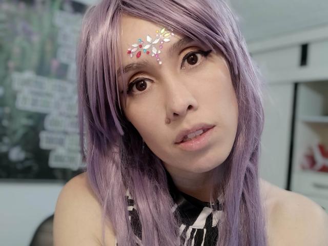 AnnaBecovka with purple hair, face glitter, and brown eyes, looking at the camera with slightly parted pink lips. She's wearing a black and white striped top.