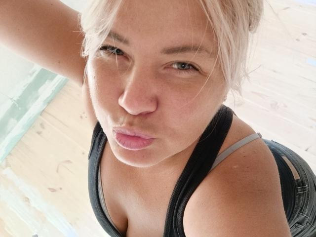 CurlyDreams with puffy pink lips, blonde hair in a messy bun, wearing a black tank top showing small tits and a grey strap, looking up at the camera.