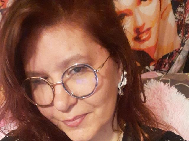 Choukette tiene la piel clara, cabello castaño y lleva gafas redondas con montura de metal. Tiene senos pequeños y está sonriendo ligeramente. Lleva pendientes blancos..