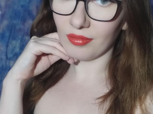 PaulinaSunny with shiny red lips, black glasses, and brown wavy hair, touching her chin, showing pale skin and small tits, against a blue background.